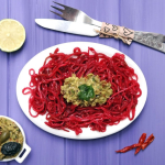 Un piatto bianco ovale contenente degli spaghetti crudisti di colore rosso, conditi con una salsa al centro. Sul piano una ciotola con altra salsa, tre peperoncini secchi, delle posate e mezzo limone. Il tavolo è di colore azzurro.